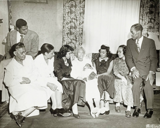 The family of Jerome Rose is seated in an indoor setting.  Everyone's attention is focused on the baby (center) except for the baby's great-grandmother (Barbara Jones) who holds her.  The women are dressed in long dresses and both of the men are in suit and tie. Back: Left-Jerome Rose Seated: L-R: Mother McBride, Mother Jennie V. Boyd, Barbara Rose,  Mother Jones (holding infant, Jerome Rose's grandmother), Betty Duncan (Rose), Carolyn Body, Eld Boyd.