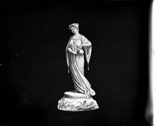 Close-up view of a statue that depicts a woman who pours liquid from a pitcher into a cup. She wears a robe.