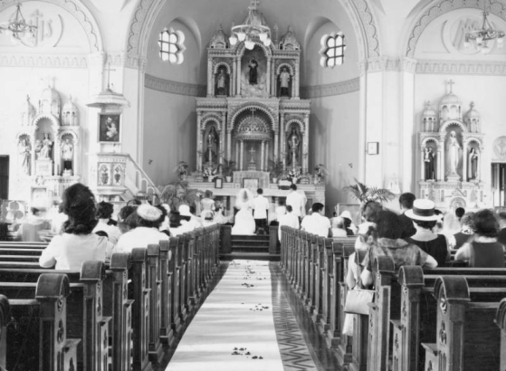 Interior view of the wedding of Lupe Morales and Gene Vigil at St. Cajetan's Catholic Church, 1190 Ninth (9th) Street, in the Auraria neighborhood, Denver, Colorado. The couple kneel in front of the altar, shows religious paintings and statues. People are in the pews, and rose petals are sprinkled along the aisle.