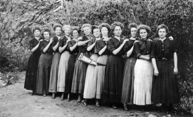 Twelve young women stand in a line near a leafy bush and a rock wall in Eldorado Springs, Colorado.  Each of the women has her right hand on the shoulder of the girl in front of her. A pennant flag with "Eldorado Spgs.," printed on it hangs from the waist belt of a girl.