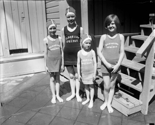 Four young girls stand in their swim apparel at the resort in Eldorado Springs, Colorado. Three of the girls wear swim caps, and their bathing suits has "Eldorado Springs," printed on the top section.