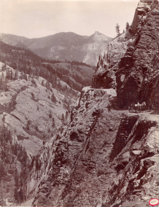 A driver in a horse-drawn buggy and a man on horseback stop on the Otto Mears toll road on a high cliff probably Ouray County, Colorado. Shows talus and rock formations, the narrow road is buttressed with logs. A passenger sits by the road.