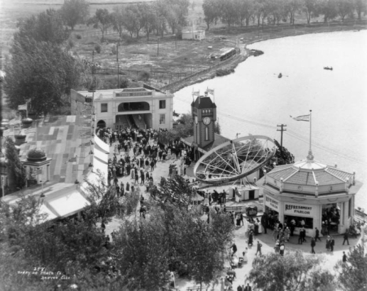 People walk by a stage, clock tower, mechanical ride, Lake Rhoda, and a concession stand with sign: "Refreshment Parlor" at Lakeside Amusement Park in Lakeside, Colorado.