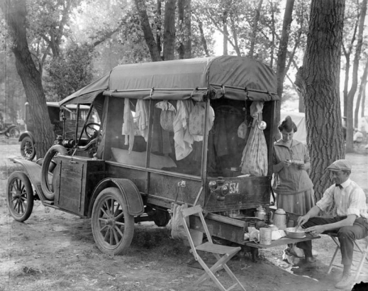 A man and woman prepare a meal in the back of a camper at Rocky Mountain Lake Park auto camp (motor camp) in the Berkeley neighborhood of Denver, Colorado. The grill, coffee pot, pans, tins and other utensils sit on a drawer that slides under the car. Clothing and bags are hung on the inside of the camper and are exposed by canvas flaps that are rolled up.  The windows are covered with screen.