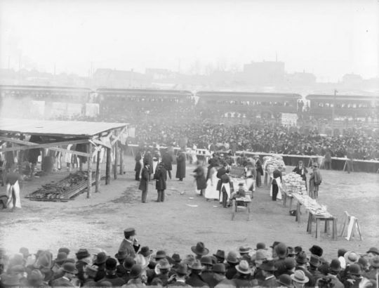 Men in aprons prepare and organize food for a barbecue at the Denver Union Stock Yard in Denver, Colorado. A table is stacked high with loaves of bread. Sides of beef are laid out over a coal filled pit. Thousands of people crowd outside barricades. Barrels of beer and tables with pitchers are set up along the barricades. A Passenger train is near by. Play bills reading: "Shall We Forgive Her?" are near the train.