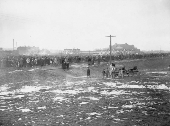 Thousands of people crowd an open area near the Denver Union Stock Yard in Denver, Colorado.  People stand along the railroad tracks, and on roofs in the distance.  Several men stand near carriages. Signs in the distance read: "Welcome, Denver Union Stock Yards," "Hotel."  Patches of snow are on the ground.