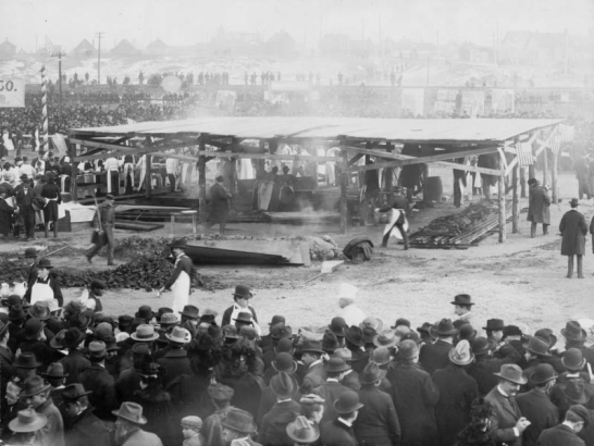 Men and women prepare food for a barbecue at the Denver Union Stock Yards in Denver, Colorado.  Three women work under a cook shed. Sides of beef are laid out over coal pits. Men in aprons stand at tables near the covered cook shed. Thousands of people crowd the area. Play bills reading "Shall We Forgive Her?" are near railroad tracks. Houses are in the distance.