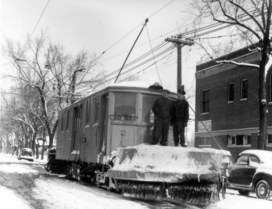 Men ride on the Denver Tramway Corporation snowplow number 2 on Caithness Place in the Highland neighborhood of Denver, Colorado. An automobile is parked nearby.