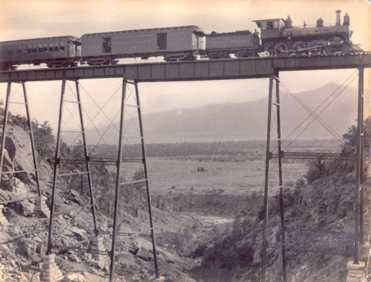 View of a Colorado Midland locomotive, baggage and mail car, and excursion car high on a railroad trestle in a gorge near Buena Vista, Chaffee County, Colorado. Shows employees of the rail company on the train. A wood frame building is in the valley