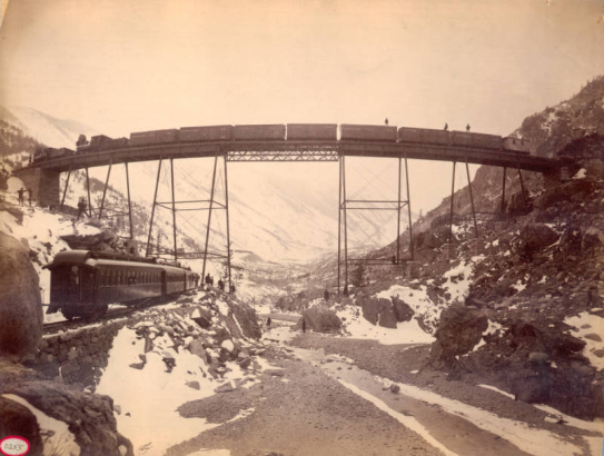 View of a Colorado Union Pacific, Denver & Gulf Railroad freight train on the narrow gauge High Bridge trestle near "The Loop," a route over Clear Creek near Georgetown (Clear Creek County), Colorado, men stand on top of the freight cars. A passenger train is on a rock rail embankment below the bridge, passengers and rail workers stand on rocks near the creek; snow-covered mountains are in the distance.