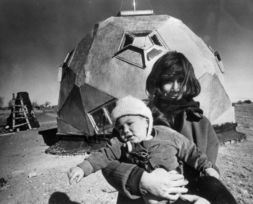 A woman holds a child bundled in a coat, hat, and mittens in Drop  City, Colorado, in Las Animas County.  They stand outside of a geometrically-shaped dwelling (geodome) with pentagon-shaped windows.  A coarse, wood ladder leans against a conical-shaped structure in the background.