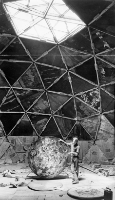A man stands in the interior of a partially-finished geodesic dome in Drop City, Colorado, in Las Animas County.  Light streams through the geometrically-shaped window at the top of the structure.  The man holds a circular painting with his right hand and stands amidst other pieces of art on the floor.