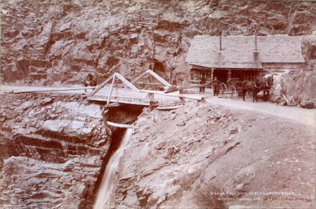 Men pose near a toll gate on the Otto Mears toll road in Ouray County, Colorado. A horse-drawn buggy, wood frame building, and a man on horseback are nearby. Bear Creek Falls is under the bridge.