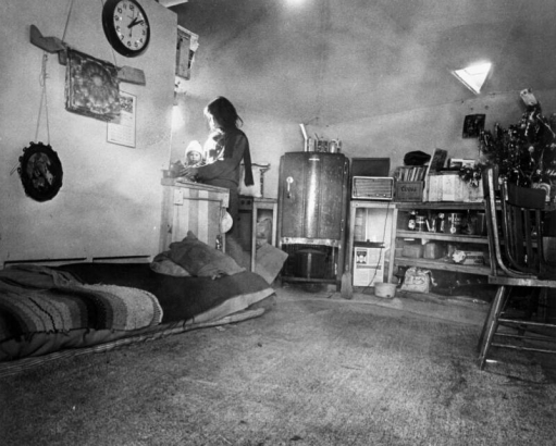 Interior of a geodome in the community of Drop City, Colorado, in  Las Animas County; shows bed covered with blankets on floor, kitchen area including refrigerator and sink, a clock on wall set to 1:10, and light through triangular-shaped window on ceiling.