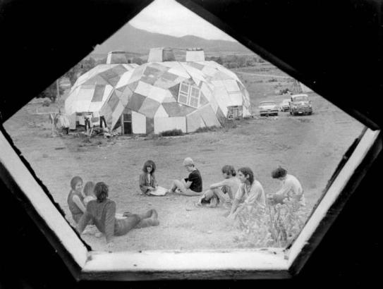 View taken from inside a geodesic dome looking outside at people sitting on the ground in Drop City, Colorado, in Las Animas County.  Three geodesic dwellings are in the background and have cars parked next to them.