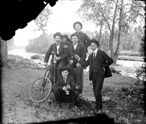 Outdoor portrait of men with a bicycle and bottles of beer possibly near the South Platte River in Denver, Colorado. They wear suits, bowties, and wide-brimmed hats.
