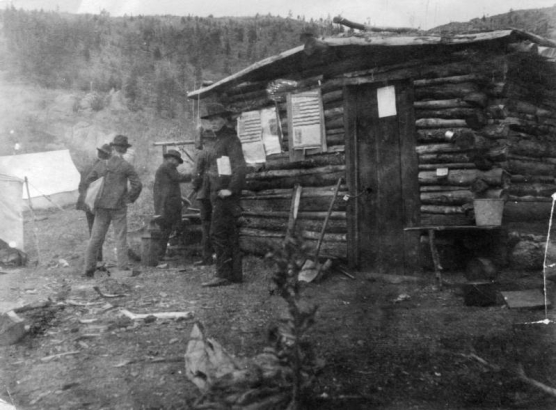 Five miners stand outside of a log cabin in Dunnville, Colorado.  Tents are in the background and debris and garbage litter the ground. Posters are attached to the side of the cabin.