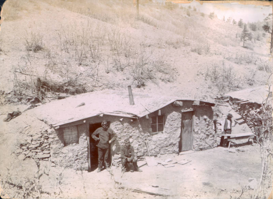 Miners pose near mud plastered dugout houses (a duplex) in Colorado. they wear hats with miners lights. One man smokes a pipe.