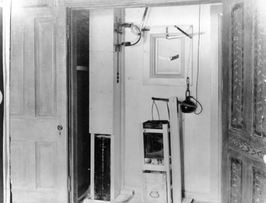 Levers, switches, and a pulley with a weight and a square can are part of the automatic hanging machine at the State Penitentiary in Canon City, Colorado. Wood molding frames a slotted window in the wall, and doors flank the apparatus. 
