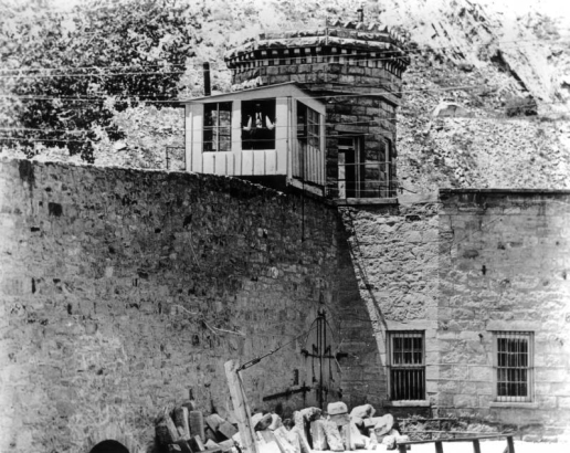 Stone walls at the State Penitentiary in Canon City, Colorado, have barred sash windows and a gate with iron fasteners. A frame guard shack with a stovepipe tops the wall; a man looks from the window. A faceted stone tower with sash windows and a cornice is behind, at the corner. Rough cut stone is piled at the base of the wall, and a beam with a turnbuckle and cable is in the foreground. Power lines cross the scene, and rock escarpments are in the background.