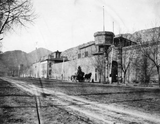 Stone walls of the State Penitentiary in Canon City, Colorado, line the sidewalk. A horse drawn water wagon on the dirt street is in front of a faceted tower with a balcony. A stone gate next to it is carved with entablature, and the original administration building is set back.