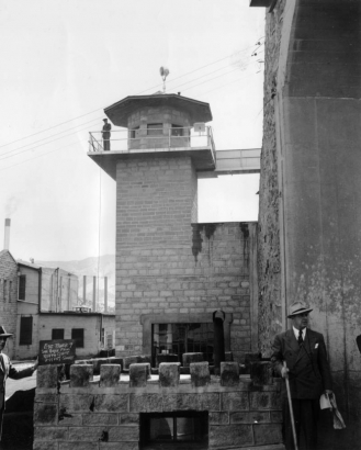 Pipe rails edge the balcony and catwalk of This guard tower at the State Penitentiary in Canon City, Colorado. Of stone masonry with extruded joints, the square tower with faceted roof has a searchlight atop it. A uniformed man at the rail lowers a rope with keys. Suited men in the foreground flank a low wall with battlements; one holds a stick and papers, the other holds a chalked sign.