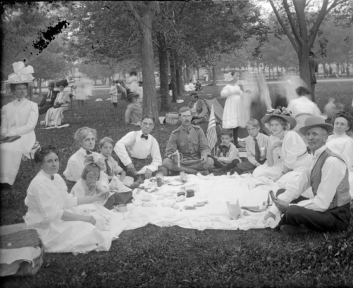 Outdoor portrait of men, women, girls, boys, and teenagers at a picnic in a Denver, Colorado park; costume includes a large hats with flowers. A man in military uniform eats a banana; a boy holds a a United States flag.