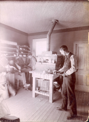 Assayers work in a lab probably in Colorado. A stove, mortars, pestles, tongs, and other equipment are in the room.