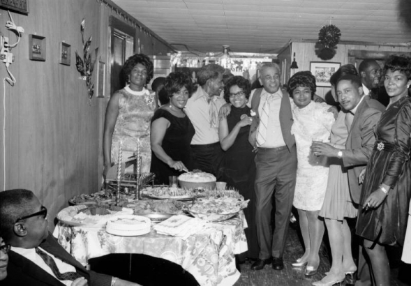 View of an Astro Jets Social Club function. A crowd of African American (Black) club members gather around a table with hors d'oeurvres at a home in Denver, Colorado. The smiling party goers pose with their arms around each other. One man wears a scarf tied around his neck. Christmas decorations hang from the ceiling.