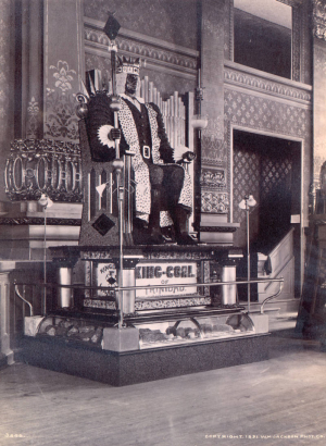 View of the statue of King-Coal against a backdrop of gilded plasterwork and ornate wallpaper in the Colorado Mineral Palace, Pueblo (Pueblo County), Colorado. The statue is made of coal, diamonds, and diamond dust, the figure is seated on a pedestal of copper and nickel with a glass display case below. Illumination is supplied by four dual-bulb floor lamps, one at each corner of the pedestal. A sign reads: "King-Coal of Trinidad." Shows a stairway to an upper floor through an archway.