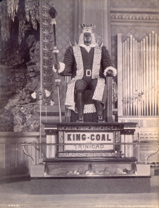 Close-up view of the statue of King-Coal in the Colorado Mineral Palace, Pueblo (Pueblo County), Colorado. The statue is made of coal, diamonds, and diamond dust, the figure is seated on a pedestal with a glass display case below. Lighting is supplied by four dual-bulb floor lamps, one at each corner of the pedestal. A sign reads: "King-Coal of Trinidad." Shows part of the stage with stalactite and stalagmite decoration. A pipe organ is nearby with a sign that reads: "The Pueblo Smelting [?]"