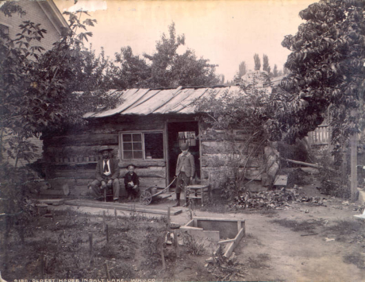 View of the oldest house in Salt Lake City (Salt Lake County), Utah; shows a one story hewn log cabin with chinking and a hewn board roof, a double horizontal window with panes of glass, a door and stone chimney. A man and two young boys stand and sit on chairs and benches near the house. Shows a small dirt yard with garden, a cold frame, a wooden push cart with one wheel, a wooden gate, trees. Shows part of a two story brick masonry house.