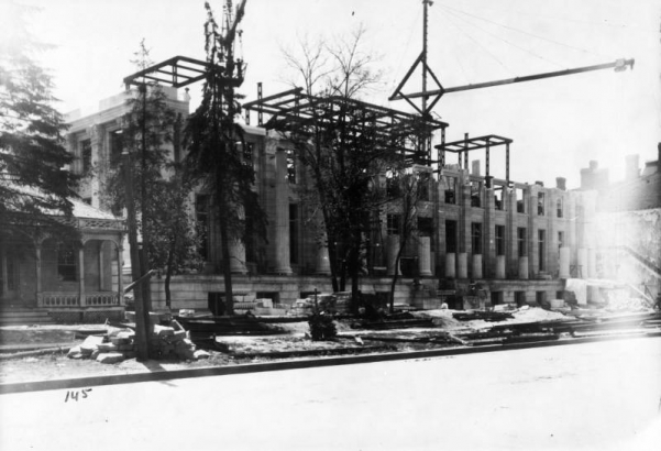 View of the partially constructed Denver Public Library at Colfax Avenue and Bannock Street in the Civic Center neighborhood of Denver, Colorado. Cranes and metal beams are on the building. The Alida Burton house and La Veta Place are nearby.