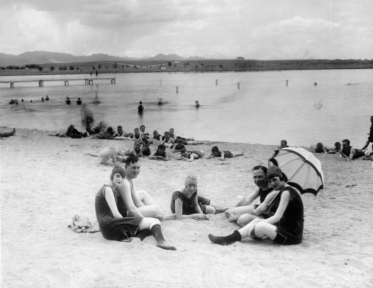 Young men and women sit on the beach at Berkeley Park in Denver, Colorado. The women wear bathing suits, bathing caps, and one woman rests a parasol over her shoulder. The men and boys nearby also wear bathing suits. Swimmers are in Berkeley Lake and on a wooden pier. The Front Range of the Rocky Mountains are is the distance.