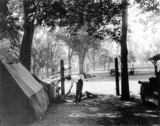 A man and woman arrange logs for a fire at the City Park automobile (auto) camp for tourists in City Park, Denver, Colorado. The woman holds a pot lid and prepares to cook.  Pots and pans hang from a fence post and a canvas tent is set up nearby. Elk lay in a fenced enclosure in the distance.