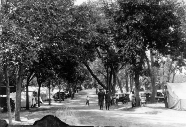 Two women walk along a road at City Park automobile (auto) camp in Denver, Colorado. Men women and children sit and stand outside canvas tents and in cars.  A banner outside one tent reads: "MO, State Fair."