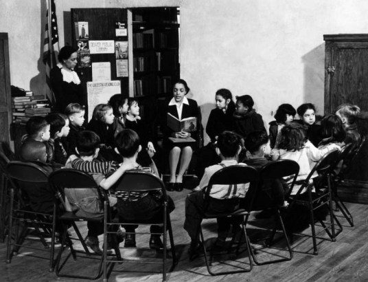 Interior view at the Community Vocational Center at the Cosmopolitan Branch of the Denver Public Library in Denver, Colorado; children listen to a Black (African American) woman read a story. Sign reads: "The Gold Star Reading Club."