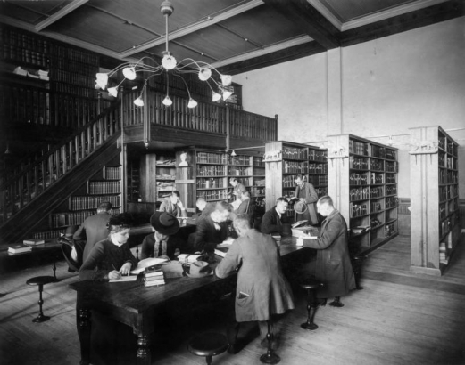 Interior view of the Denver Public Library at Denver High School in Denver, Colorado; men and women study. A stairway and hanging lamp are in the background; sculptures of lions adorn the bookshelves.