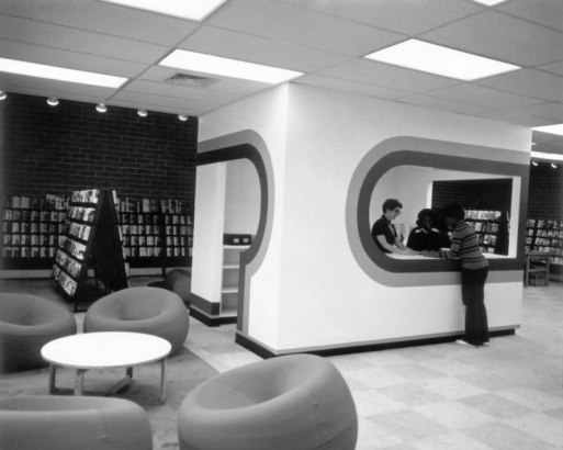 Interior view of the Five Points Branch of the Denver Public Library in Denver, Colorado; shows "mod" decor, wall graphics, curved wall cutouts, and amorphous chairs. A Black (African American) staff member helps a woman.