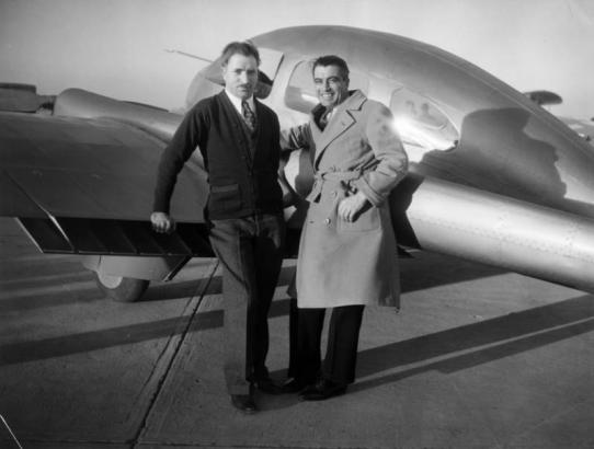 Portrait of Tom Sheldon (aircraft designer) and Raymond Wilson (in spotted tie), pilot, educator, and businessman. They pose by a small passenger airplane.