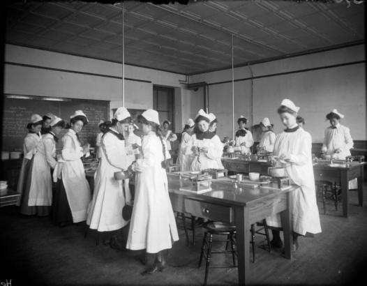 Girls in smocks and caps stir food and peel apples in a cooking class at Mitchell School in Denver, Colorado. Shows a coffered ceiling, long wooden tables with burners, and recipes on the blackboard.