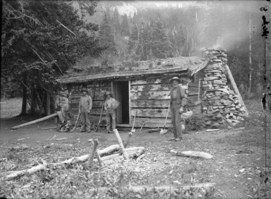 Men pose by a miner's cabin identified as Hepburn's cabin in Miners Basin in the La Sal Mountains (Grand County), Utah. Shows mining tools, ropes and a rifle. The log cabin has a stone chimney and a sod roof.