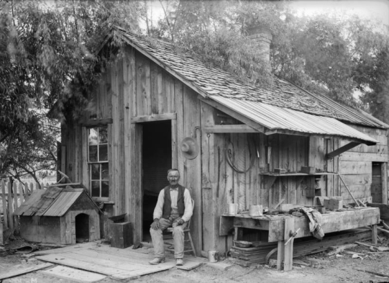 Hughy, a man in cowboy boots and a vest, holds a pipe and sits in front of a wood cabin with a wooden shingle roof in Colorado or Utah. A wooden doghouse is by the porch. Metal buckets, a branding iron, a drill bit, bundles of wire, hats, horseshoes, and a workbench are near the cabin.