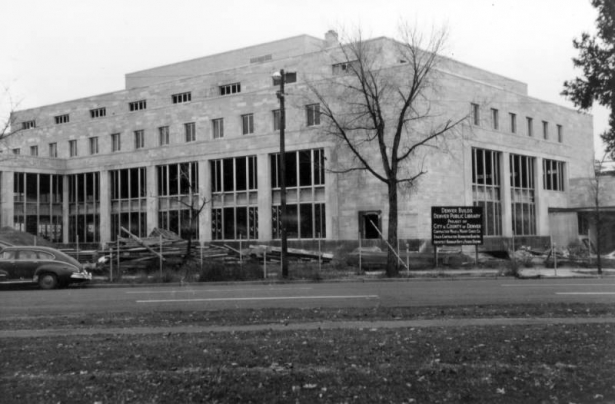 View of Denver Public Library construction at 1357 Broadway Street in the Civic Center neighborhood of Denver, Colorado; shows stone walls, window openings, a parked car, and building materials. Sign reads: "Denver Builds Denver Public Library Contractor Mead & Mount Const. Co. Stack Contractor Remington-Rand Inc. Architect Burnham Hoyt & Fisher Denver."