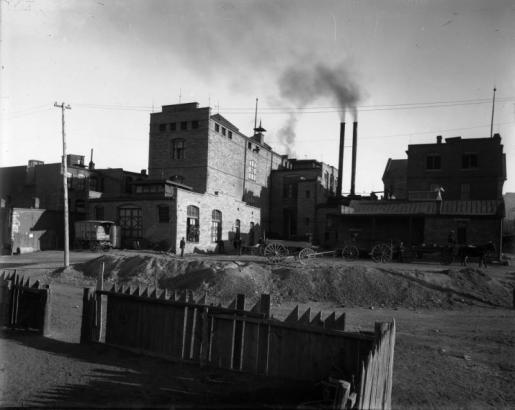 View of the Schneider Brewing Company in Trinidad (Las Animas County), Colorado. Men sit on and stand near a horse-drawn wagon parked near the brick buildings.