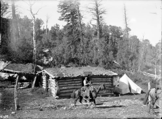 Two men on horseback and a third man stand in front of Geyser cabin in the La Sal Mountains, probably San Juan County, Utah. Signs read, "Geyser cabin," "La Sal and Geyser Express Office," and "Stage Office." Shows a tent beside the cabin and a carcass hanging from a corner of the cabin.