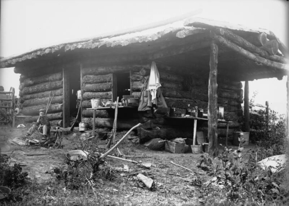 A boy sits in front of Buckeye Cabin in the La Sal Mountains, Utah. A sign reads, "Assay Office Buckeye Cabin." On a table are reagent bottles, a box of assay weights, a beaker, test tubes, glass-stoppered acid bottles, evaporating dishes, and a hot air sterilizer. Also shows a rifle, a shotgun, a gold washing pan, a pickaxe, a hand pick, and buckets.
