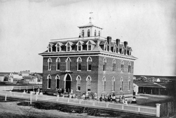 Young women stand in the fenced yard of Wolfe Hall School in Denver, Colorado. The brick building has a mansard roof, dormers, and cresting. A woman stands on the flat roof near a cupola. A cross is mounted on the top of a building. Houses and outbuildings are in the distance.