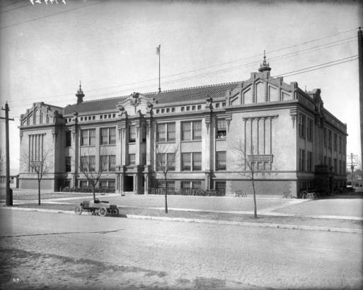 A group of children look out a second story window at Gove Junior High School at East 14th (Fourteenth) Avenue and Colorado Boulevard in Denver, Colorado. The building is a two and one-half story brick structure with carved stone decoration. Bicycles are lined up in bike racks, and a car is parked in the street.