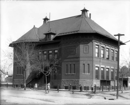 Children stand in the yard of Ironton School at East 36th (Thirty-sixth) Avenue and Delgany Street in the Five Points neighborhood of Denver, Colorado. The school is a two story brick structure with a high pitched, sloping roof.